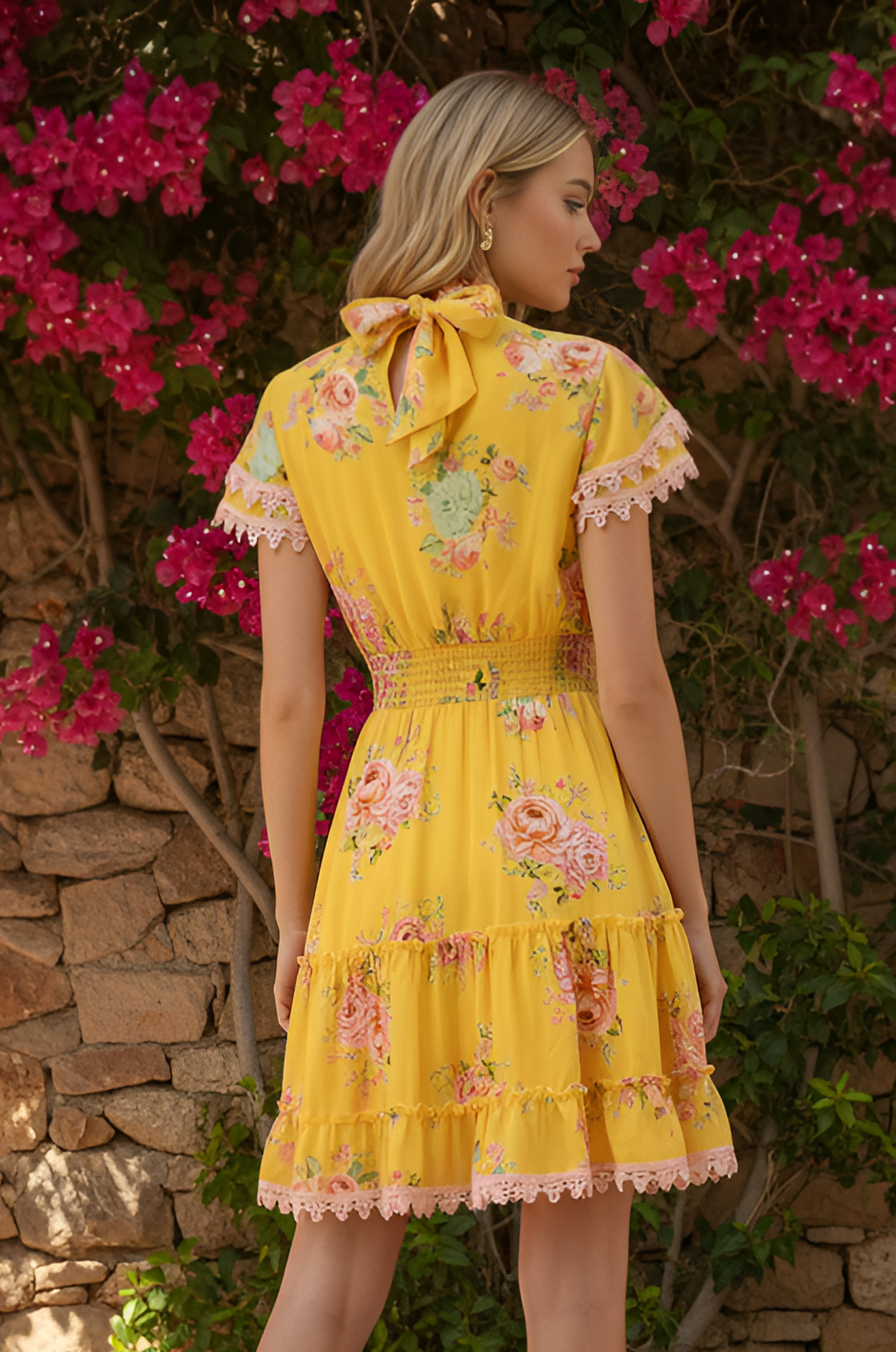 A woman stands outdoors by a stone wall and pink flowers, seen from the back wearing the Maya Sunshine Yellow Short Floral Casual Dress.